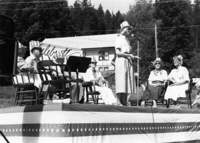 A woman speaking on a stage during Bovill's Statehood Day celebration, while others sit behind her.