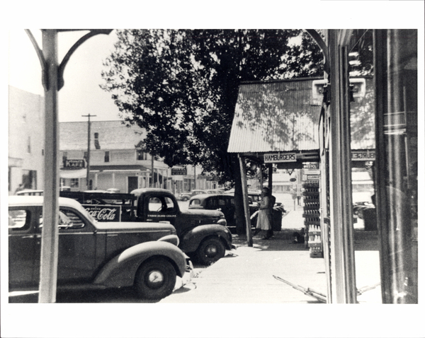 Sidewalk with a view of a hamburger place, Korner cafe, and some old cars parked on the side. Image contains the text: "KAFE Coca Cola HAMBURGERS"