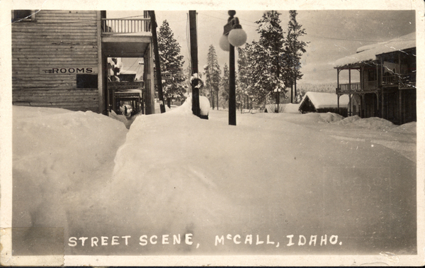 McCall in the snow, in view there is buildings, a light post, and snow covered pine trees. Image contains the text: "ROOMS  STREET SCENE, MCCALL, IDAHO."