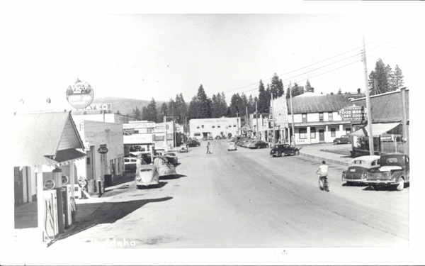 New Hotel McCall in the background, in view are several buildings like motor oil shops a food market, on the road someone riding a bike, to the sides of the road several parked cards. Image contains the text: "Here is the extracted text from the image:  Vico Motor Oil Associated Westcott Oil Co  Hotel McCall  Stockgill's Food Market Dry Goods Gifts - Locker Cafe"