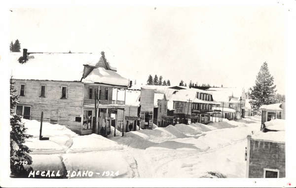 Snow covered street view of Main St., in view there are several large buildings like the Geelan’s Dept. Store, Drug store, ice cream parlor, and more. Image contains the text: "MCCALL IDAHO 1924  ROOMS  LES DRUG  CEELINGS DEPARTMENT STORE"