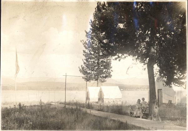 View of three tent-frame cabins, two people sitting on a picnic table, and a path leading down to the lake