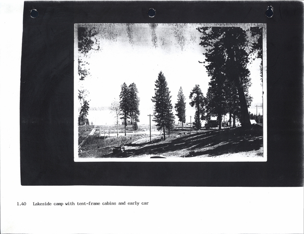 View of several large pine trees, a early car driving on the road, and the lake in the background. Image contains the text: "1.40 Lakeside camp with tent-frame cabins and early car"