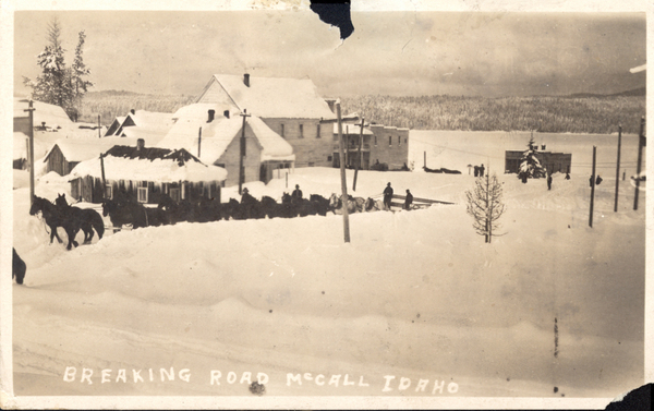 McCall’s winter, the road is being broken with a 20-horse team on 3rd Street, the view is of snow covered road, houses/buildings, and lake. Image contains the text: "BREAKING ROAD McCALL IDAHO"