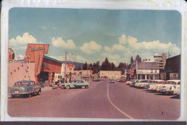 E. Lake street there are cars parked on both sides of the road, buildings are in view like The Western Store Hardware, Conoco, Hotel McCall, Lola’s Cafe, and more. Image contains the text: "Western HARDWARE Tavern HOTEL MONTANA DRUGS MEN'S SHOP"