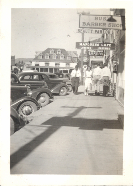 View of people walking in the sidewalk walk, there are cars parked on the left hand side of the photo, large building/ businesses like a barber shop, cafe, bakery, and more. On the back there is a handwritten note, “Picture taken in Summer 1937while I was in the CCC at the French Creek. I have other pictures of French Creek. If you use this in the Star News please send me a subscription and advice. Thank you. Rexlu Catarrelle.”. Image contains the text: "BUSS BARBER SHOP BEAUTY PARLOR MARQUESS CAFE LUNCHES PASTRIES Mary Ann's BAKERY"