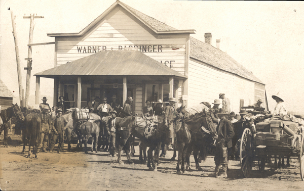 Original postcard with a view of the Lardo Warner store in the background, the foreground has several men with horses, and wagons. Image contains the text: "WARNER & BARRINGER"