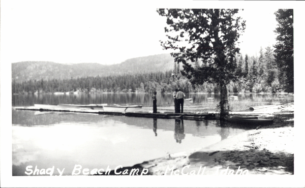 Shady beach camp shoreline, there is a dock, three men are on the dock, some boats, and in the background the mountains. Image contains the text: "Shady Beach Camp McCall Idaho"