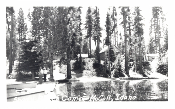 View from a dock, with a view of the shoreline, trees, three men on a dock, and cabins in the background. Image contains the text: "Shady Beach Camp McCall Idaho"