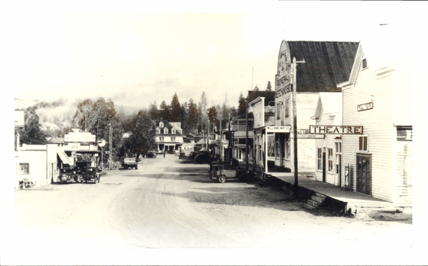 Main St., with a West view, with the Theatre, General merchandise, other buildings, and early cars parked to the sides of the road. This photo was used as notecard. Image contains the text: "THEATRE WILLIAMS MAY CO NARROW GAUGE CO RANGE NATIONAL LIGHT  SING SERVICE STANDARD OIL CO CAFE"