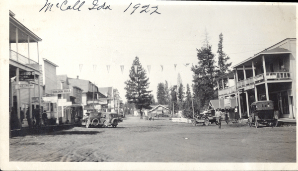 East view of Main St. in 1922 visible buildings are the Golden Rule, Drug Store, Grocery Store and more, there is the road and early cars parked on either side of the road. Image contains the text: "McCall Ida 1922  ICE CREAM  GOLDEN RULE  ROOMS"