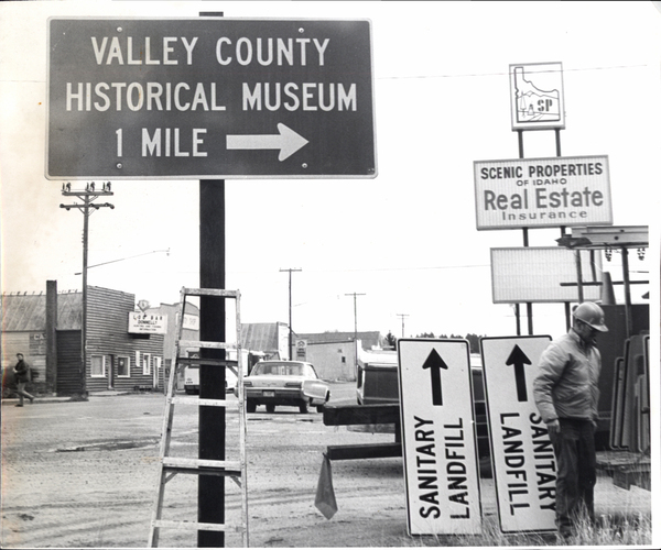 Donnelly Main St. with Log Bar. Image contains the text: "VALLEY COUNTY HISTORICAL MUSEUM 1 MILE  DONNELLS  CAFE  SCENIC PROPERTIES OF IDAHO Real Estate Insurance  SANITARY LANDFILL SANITARY LANDFILL"