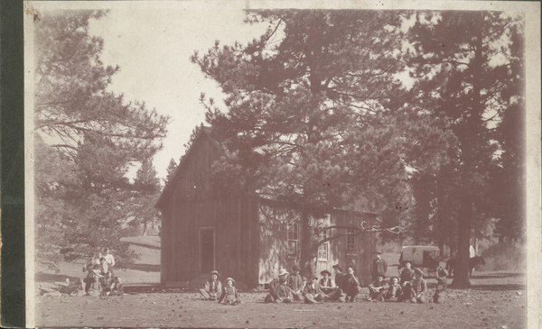 Large group of people outdoors, with board and batten cabin, horse, and enclosed buggy.