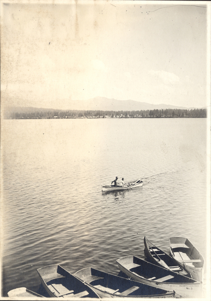 View of Payette Lake, McCall in the distance, on the lake two men rowing a boat, on the bottom several row boats stationed. Image contains the text: "PAYETTE LAKE CLUB   No. 3"