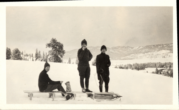 Hill with a view of the snowy mountains, there are three men in total with a sled, one of the men is sitting on the sled