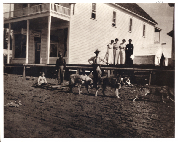 Building with a child of a sled being pulled by dogs on a dirt road, there are also two men and four women watching