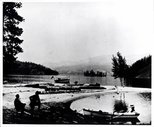 View of the beach at Payette lake, on the shore there are two men, offshore there is a row boat and the baot “Winston”