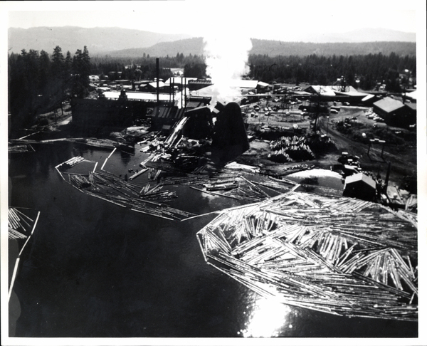 View of mill and three or four log booms, showing log slide into mill