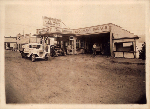 Goodman’s Garage, in view there are several men, and ealry cars in front of the building. Image contains the text: "GOODMAN'S SUPER SERVICE CAR WASH GREASING GOODMAN'S GARAGE GOODMAN'S Tourist Information PYROIL"