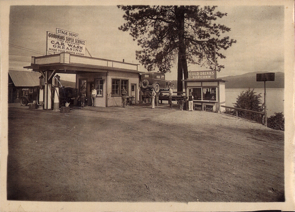 Goodman’s Super Service, there is also a small snack shack next to it, several men in view, and a car lifted. Image contains the text: "STAGE DEPOT GOODMAN'S SUPER SERVICE THE REPAIRMEN CAR WASHING GREASING  MAKE YOUR WANTS KNOWN COLD DRINKS POPCORN"