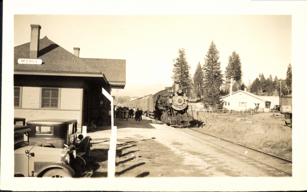 Train at McCall Depot, crowd waiting, and early cars are also in view. Image contains the text: "MCALL"