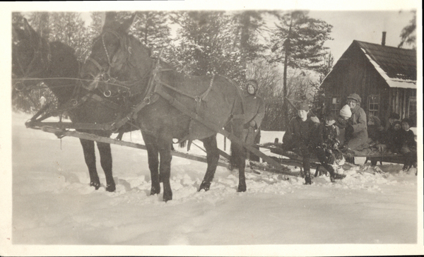 A two mule team with sled and passengers, and cabin in the background