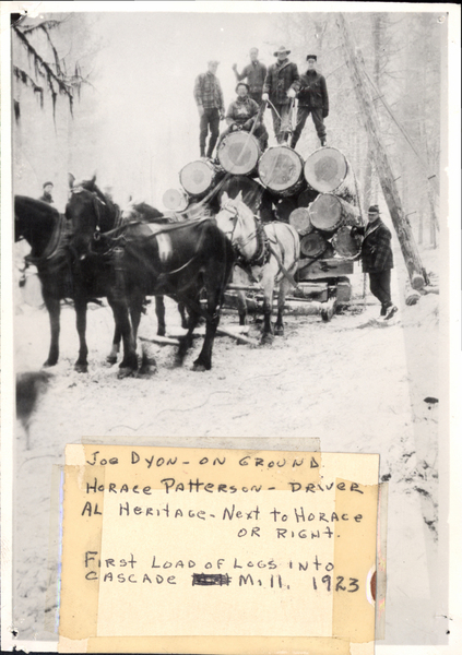 Six men with four-horse team, log load on sled, “First load load of logs into Cascade Mill 1923,” most men named, Joe Dyon- on ground, Horace Patterson-Driver, Al Herotage-next to Horace or right.. Image contains the text: "JOE DYON-ON GROUND   HORACE PATTERSON-DRIVER   AL HERITAGE-NEXT TO HORACE OR RIGHT   FIRST LOAD OF LOGS INTO CASCADE M Ill 1923"