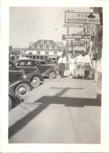 View of people walking in the sidewalk walk, there are cars parked on the left hand side of the photo, large building/ businesses like a barber shop, cafe, bakery, and more. On the back there is a handwritten note, ���Picture taken in Summer 1937while I was in the CCC at the French Creek. I have other pictures of French Creek. If you use this in the Star News please send me a subscription and advice. Thank you. Rexlu Catarrelle.���. Image contains the text: "BUSS BARBER SHOP BEAUTY PARLOR MARQUESS CAFE LUNCHES PASTRIES Mary Ann's BAKERY"