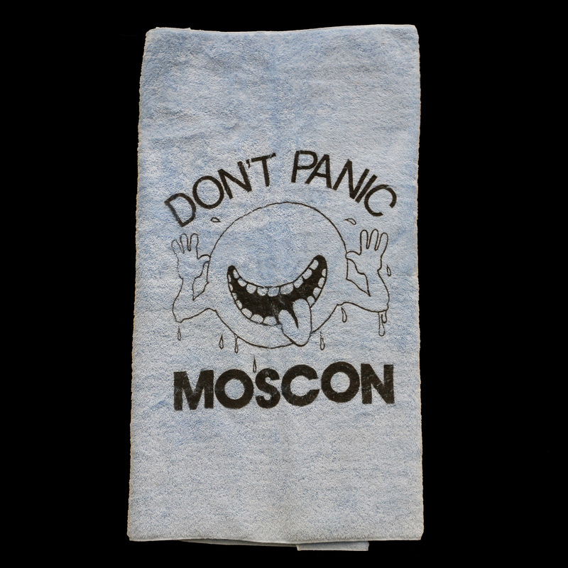 A large, blue towel with text reading "DON'T PANIC MOSCON" and an image of "The Cosmic Cutie" from Douglas Adams' The Hitchhiker's Guide to the Universe. An image of Leonard Nimoy holding this towel up at MosCon V can be found on page 10 of the MosCon VI booklet.