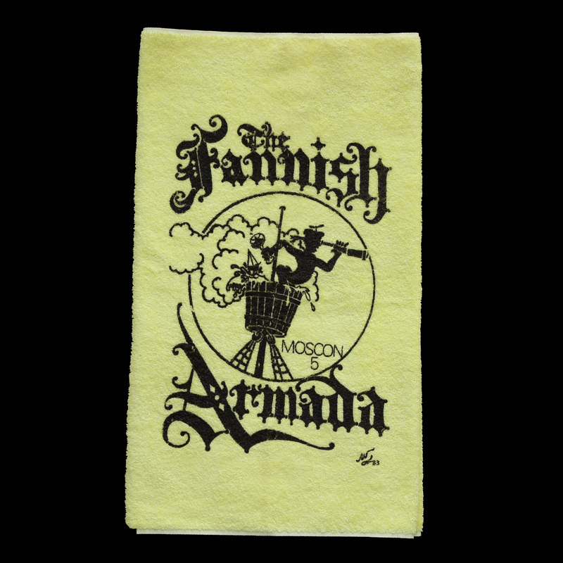 A yellow towel with a circle bordering an image of a sailor in a crow's nest holding a hand held telescope with clouds in the background. The clouds pass out of the border slightly and the text "Moscon 5" is in the lower right corner of the circle. The image separates the bold, black, scrawly text stating "The Fannish Armada," with "The Fannish" on top and "Armada" on the bottom. 