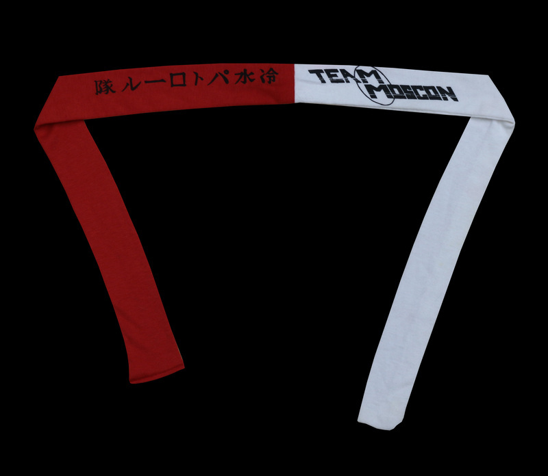 A red and white cloth headband that reads "Team MosCon" in English and "Cold Water Patrol" in Japanese. From the MosCon VII (1985) booklet, p. 15: "Overflowing as we are with new and innovative ideas, we actually accomplished one this year. Instead of the standard t-shirt or towel, we have, in limited supply, Team MosCon headbands. We chose headbands in part to provide our membership with a handy place to attach their nametags while in the jacuzzi, and also as a method of identifying the Con-com. (Ours are blue.) The idea was inspired by the film, Buckaroo Banzai. The Team MosCon logo was designed by Jacqualynn Duram and the Japanese was caligraphed by Ellen Thisted. The Japanese is pronounced, "Tsumetai mizu patororu-tai" and means "Cold Water Patrol." Get one quick. There will be a limited number of them for sale at the registration table."