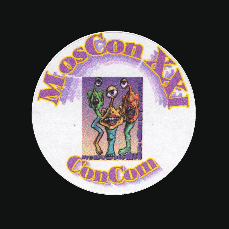 A white, circular sticker with pink text reading "MosCon XXI ConCom" and an image of 3 one-eyed alien characters standing together.