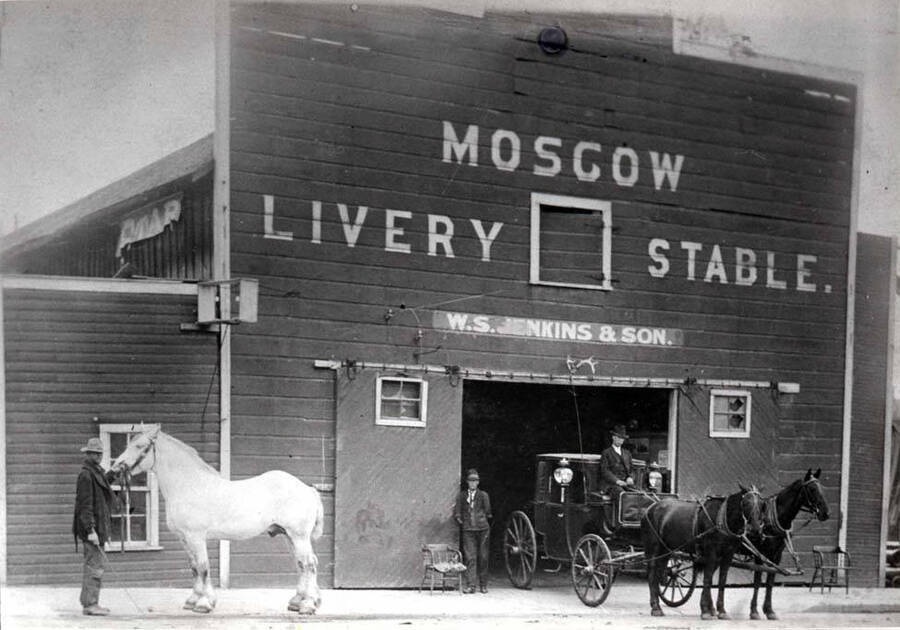 Moscow Livery Stable located at the southeast corner of Jackson and Fourth streets. W.S. Jenkins and son owners at this time. Homer Lyons with white stallion, Ernest (Short Arm) Weeks standing in doorway. Man on cab not identified. Picture from Leo Nelson of Moscow.