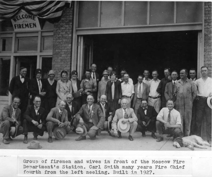 Group of firemen and wives in front of the Moscow Fire Department's Station. Carl Smith, many years Fire Chief, fourth from the left kneeling. [Building] built in 1927.