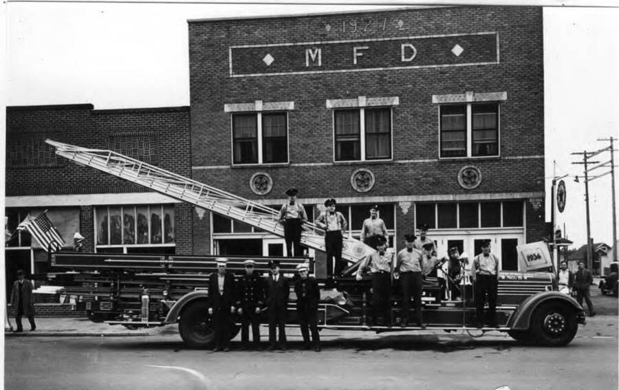 Left to right on hook and ladder truck: Wilson Rogers, Russ Tate, Ardie Driscoll, Jim Phelps, Loyd Randall. On running board: ?, Cyril Fitsimmons, Walt Schumacher. On street: Dave Millilerd, Chief Carl Smith, salesman, Harry Frazier. Volunteer Fire Department showing new fully-equipped Seagrave Junior aerial ladder truck. About 1937.