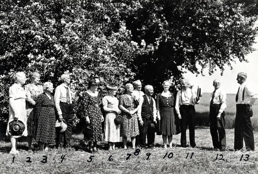 Pioneers in front of the poplar tree planted May 6, 1871, by Mr. and Mrs. George W. Tomer. These pioneers were among the many people and students in the tour Tracing The Old Nez Perces Trail, sponsored by Dr. C.J. Brosnan, head of the department of American History, University of Idaho. This was during summer school in 1937. No. 1- Maude (Barton) Hunter, 2- Ruth (Naylor) Perkins, 3- Margaret Mills, 4- George Northrup, 5- Mrs. Emma Clyde, 6- Mrs. Daniel Gamble, 7- Nellie Summerfield, 8- Gainford P. (Gub) Mix, 9- Walter McClintic, 10-Belle (Estes) Carssow, 11- Charles J. Munson, 12- L.G. Peterson, 13- George M. Tomer (son of George W. Tomer) was the official guide for the tour.
