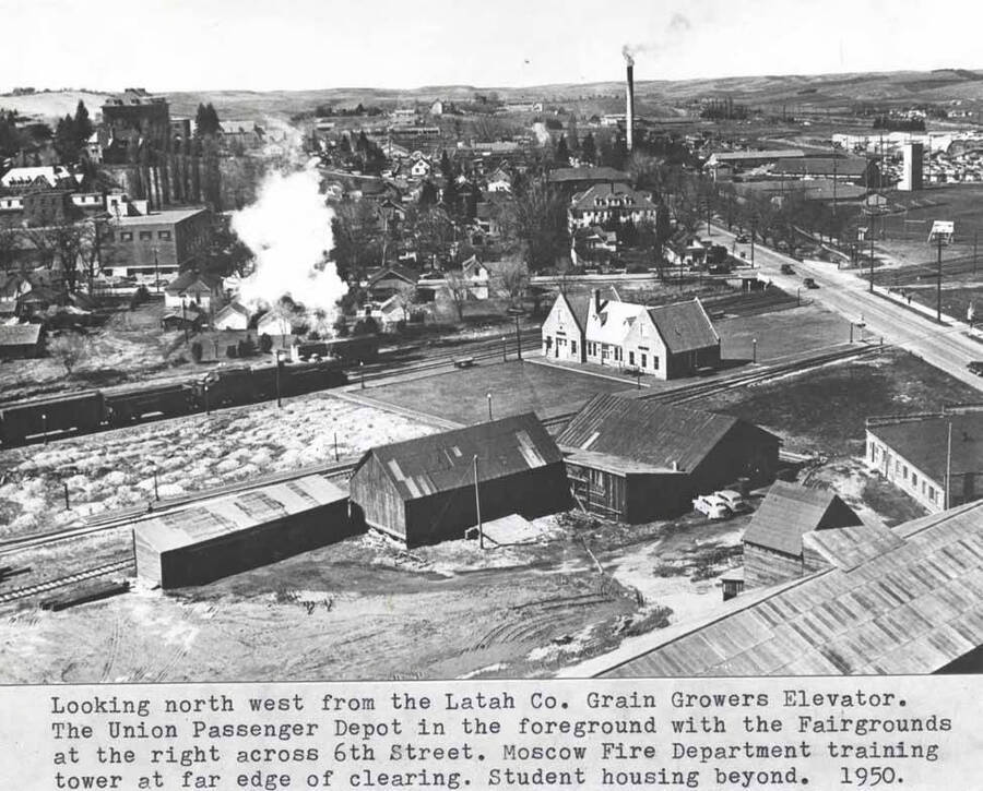 The Union Pacific Railroad passenger depot in the foreground with the fairgrounds at the right across Sixth Street. Moscow Fire Department training tower at far edge of clearing. Student housing beyond. 1950.