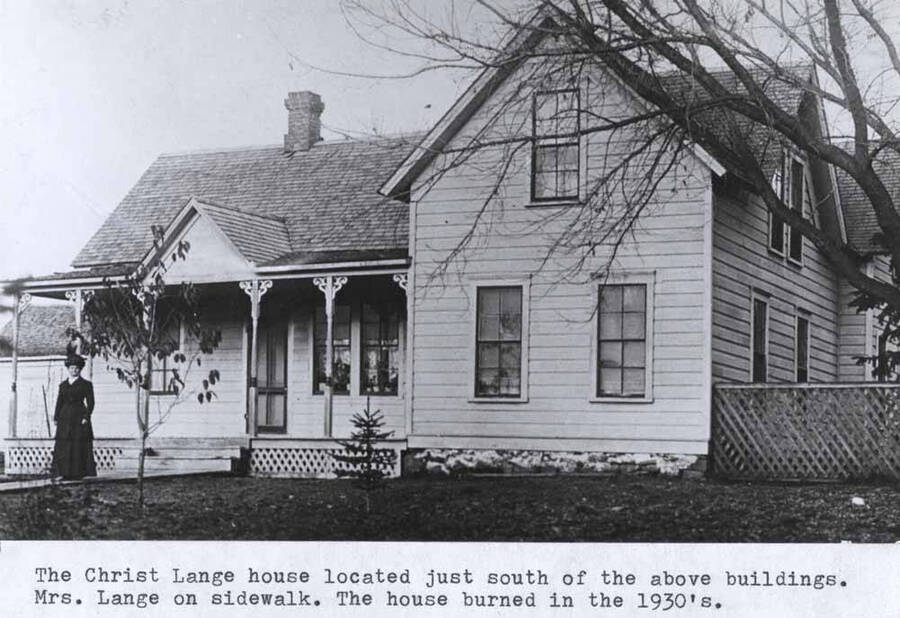 Located just south of the above buildings [90-4-027]. Mrs. Lange on sidewalk. The house burned in the 1930s.