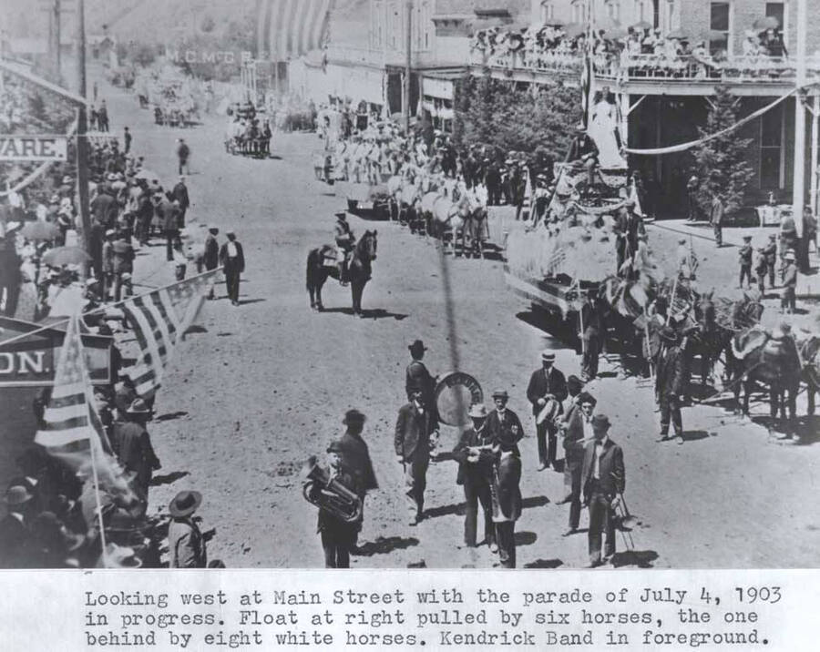 With the parade of July 4, 1903, in progress. Float at right pulled by six horses, the one behind by eight white horses. Kendrick Band in foreground.
