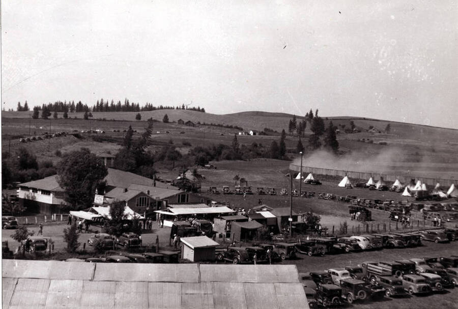 In foreground, Palouse River hidden in trees and brush with Highway 95 at the left in picture with cars parked along the roadside. See Indian teepees with dust from a race in progress beyond. Late 1930s-40.