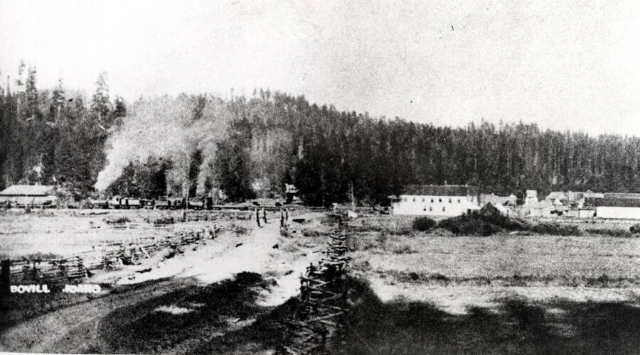 [photo of newspaper photo and caption?] Probably 1909. Note the zig-zag rail fence in the foreground. Left to right in town: warehouses, Shay locomotives (depot not yet built), park area and Bovill Hotel premises. Sherman House on Main Street, saloons at right fronting on Pine Street. In distance at the extreme right -- later location of the David boarding house -- is a frame building which was probably the recreation hall.