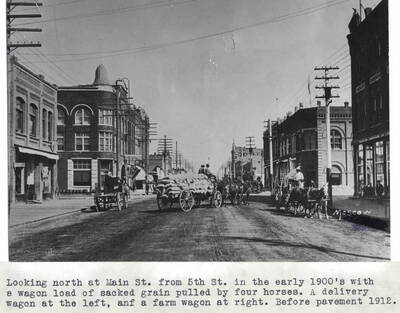 In the early 1900s with a wagon load of sacked grain pulled by four horses. A delivery wagon at the left and a farm wagon at right. Before pavement [laid in] 1912.