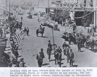 With the parade of July 4, 1903, in progress. Float at right pulled by six horses, the one behind by eight white horses. Kendrick Band in foreground.