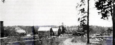 [photo of newspaper photo and caption?] Construction of Main Street in Bovill. The building at left was probably built as a store, but later housed the Major and Peters Barber Shop. The other structures are the frames of the E.K. Parker store and the Sherman House. Through the gap -- later filled by other structures, from the Bovill Drug and Spokane Hotel down to the Grant and Giles store -- are seen a Shay locomotive and tent. At right is a rod engine. Construction of the Parker store is dated by his daughter, Marie, as 1908.