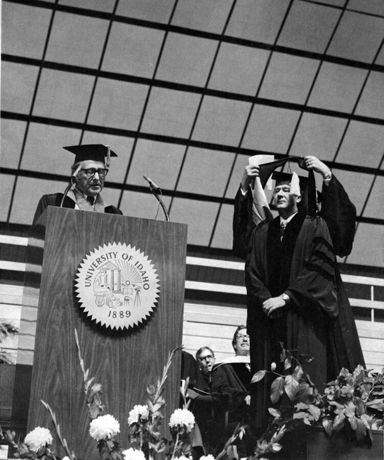 University of Idaho President Ernest Hartung awards Governor Cecil Andrus an honorary Doctor of Law degree. Andrus is hooded by an unidentified individual behind him.