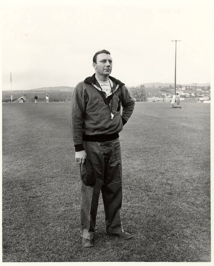 Millard F. 'Dixie' Howell, the head coach for the Vandal football team at the University of Idaho standing on the field while players move equipment behind him.