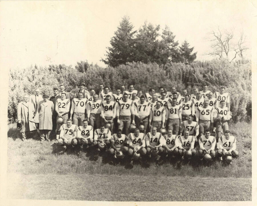 A group photo of the University of Idaho varsity football team in full gear with their coaches in front of some bushes and trees in a field.