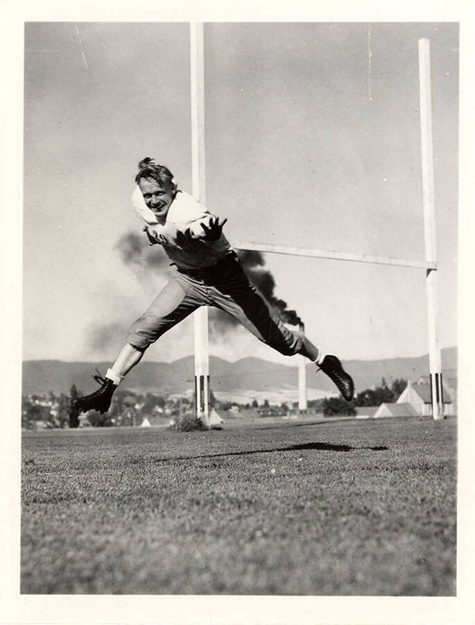 Action shot of Gordon Price, left halfback for the University of Idaho football team, running with the ball preparing for a tackle. Behind him you can observe the goal post and a smokestack  billowing black smoke.