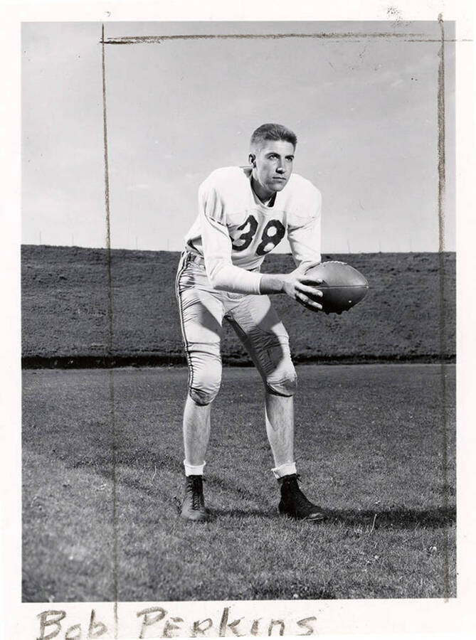Football player for the University of Idaho, Bob Perkins (#38), standing on the field and leaning over to hold a football.