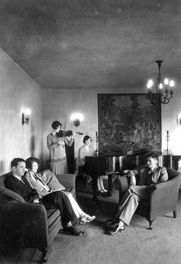 Interior view of the living room of the Kappa Alpha Theta house, which is on southwest corner of University and Deakin Avenues. Two women are playing musical instruments, while others sit around.